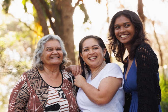 Three women of different generations standing close together and smiling looking off in the distance