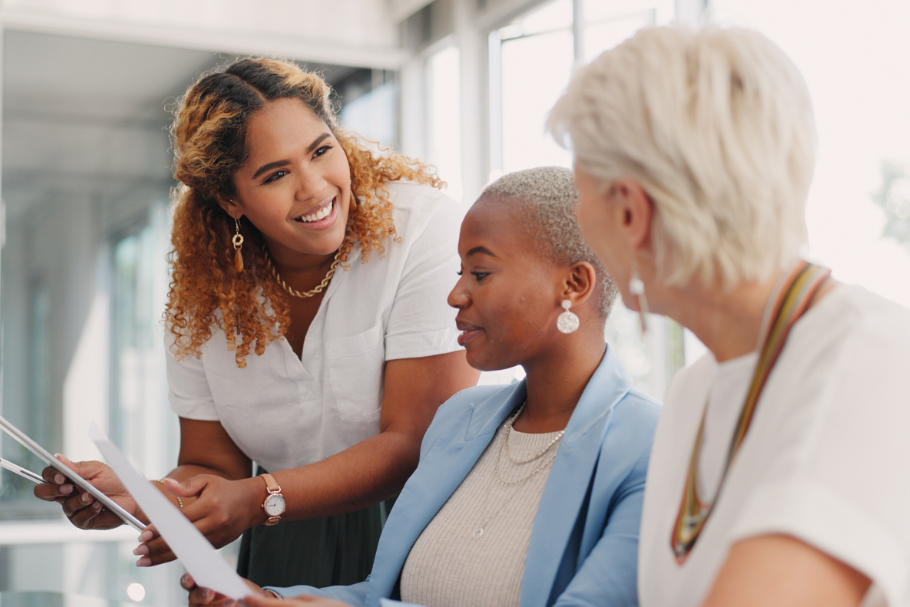 A professional is standing up and smiling while talking with two professionals sitting down reviewing paperwork in an office setting with bright, natural light coming through the windows.